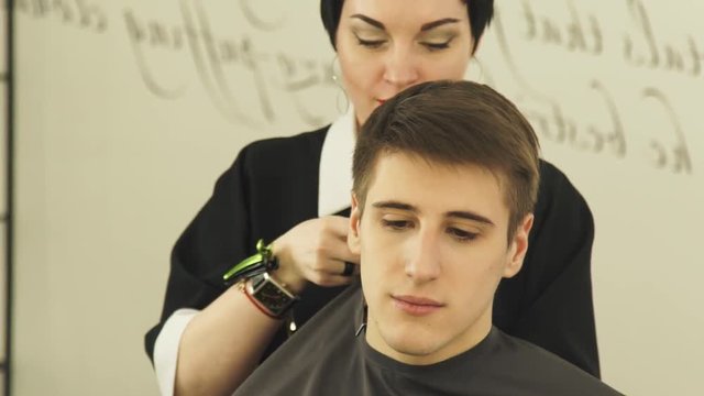 Woman hairdresser wearing cloack robe on young man before haircut in hair studio. Preparing for haircut in barber shop frong mirror.