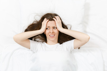 Top view of head of tired brunette young woman lying in bed with white sheet, pillow, blanket. Shocked female cover ears with hand, spending time in room. Rest, relax, good mood concept. Copy space.