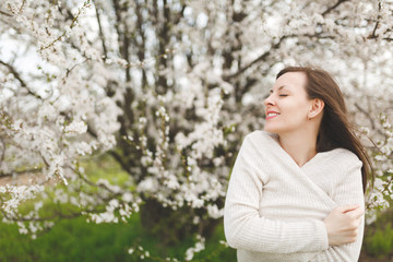 Obraz premium Young smiling relaxed tender beautiful woman with closed eyes in light casual clothes standing in city garden or park on blooming tree background. Spring nature, flowers. Lifestyle, leisure concept.