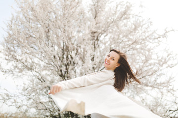 Young happy relaxed smiling beautiful woman in light casual clothes turning around standing in city garden or park on blooming tree background. Spring nature, flowers. Lifestyle, leisure concept.