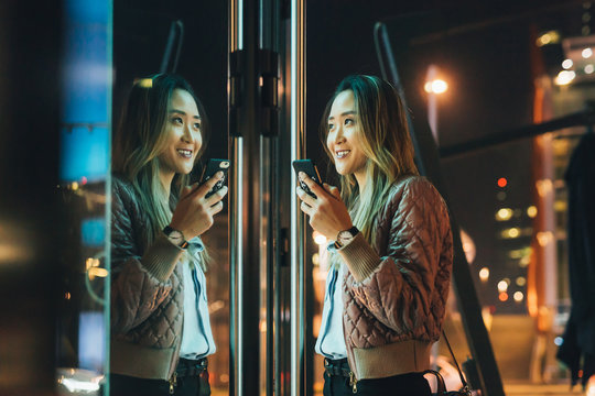 Woman standing outdoors, at night, holding smartphone, reflection in window