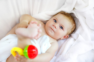 Cute little baby playing with toy rattle and own feet after taking bath. Adorable beautiful girl wrapped in white towels