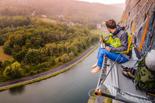 Young male rock climber sitting rock face portaledge above river Meuse, Freyr, Belgium, elevated view