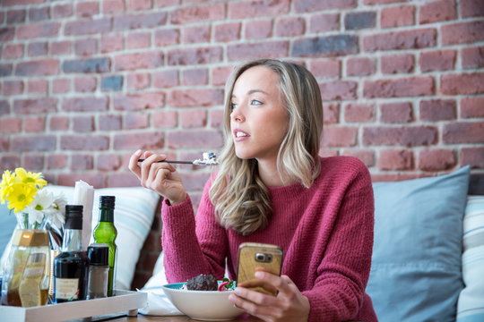 Woman sitting in cafe, eating bowl of muesli, holding smartphone