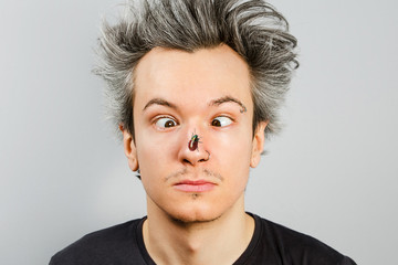 Unshorn and unshaven squint young guy with piercings on his face, with a fly on the nose, on gray background.
