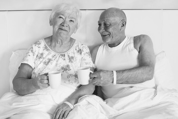 Black and white close-up portrait of handsome mature bold man in white shirt and his beautiful wife with short grey hair wearing elegant pajamas drinking morning weekend coffee together in bed.
