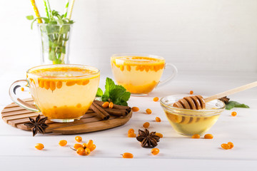Vitamin healthy sea buckthorn tea in glass cups with fresh raw sea buckthorn berries and cinnamon sticks, anise stars, mint  and honey on a white kitchen table.