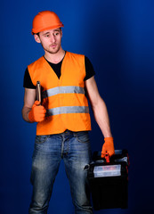 Man in helmet, hard hat holds toolbox and folder with documents, blue background. Worker, repairer, builder on dreamy face carries case with tools. Repair service concept. Repairman ready to work.