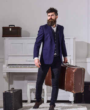 Macho stylish on strict face stands and carries big vintage suitcase. Baggage and travelling concept. Man, traveller with beard and mustache with baggage, luxury white interior background.