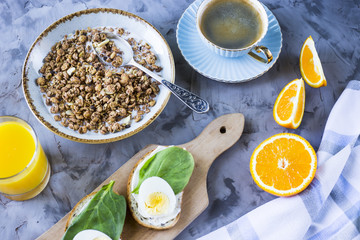 Healthy hearty breakfast - muesli, sandwich with egg, coffee, orange and juice on a gray background. Top view