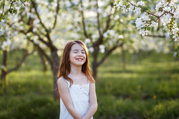 A portrait of a beautiful young girl with her eyes closed  in a white dress in the garden with apple trees blosoming having fun and enjoying smell of flowering spring garden at the sunset 