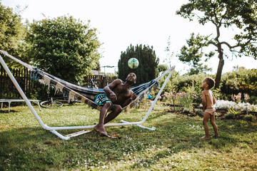 Obraz na płótnie Canvas Schwarzer Vater sitzt auf Hängematte im Garten und spielt Ball mit seinem Sohn an einem warmen Sommertag