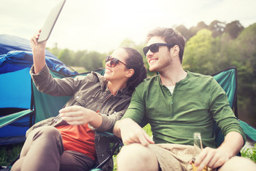 travel, hiking, technology, tourism and people concept - smiling couple with tablet pc computer sitting on chairs and taking selfie or video at camping tent