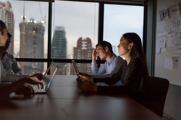 Business people team working late night in low light from laptop screen with cityscape blurred background.Overtime hours concept.