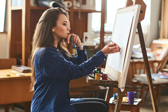 Young beautiful girl, female artist painter thinking of a new artwork idea and ready to make the first brushstroke on a wooden easel with a brush in studion interior