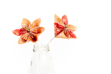 Two orange origami flowers in transparent glass bottle on white background.
