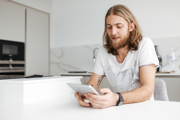 Portrait of young smiling man sitting and using digital tablet in kitchen at home isolated
