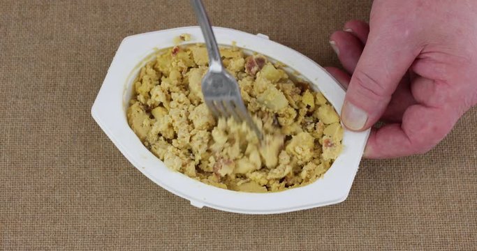 Top view of a microwaved meal of scrambled eggs with potatoes and bacon in a white plastic tray being mixed with a fork then taking a forkful at the end.