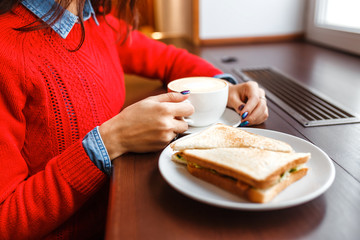 young girl student eating a sandwich and drinking coffee in a cafe near window, office dinner break concept