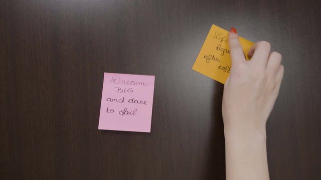 Female hands with red nails sticking various colored post it notes with motivational quotes on her dark wooden dressing to see them every day
