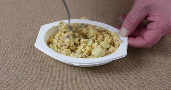 A microwaved meal of scrambled eggs with potatoes and bacon in a white plastic tray being mixed with a fork then taking a forkful at the end.