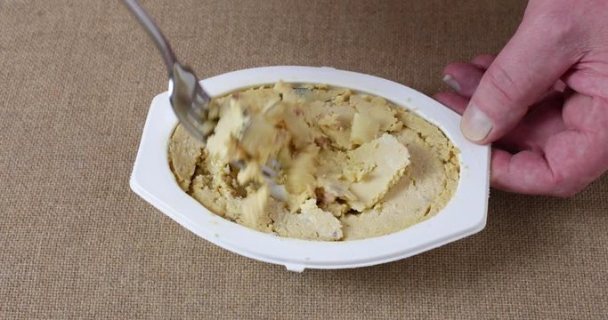 A microwaved meal of scrambled eggs with potatoes and bacon in a white plastic tray being mixed with a fork atop a brown tablecloth.