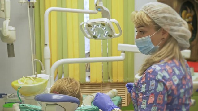 Child with a mother at a dentist's reception. The girl lies in the chair, behind her mother. The doctor works with an assistant. Procedure for drilling a tooth. Setting a temporary seal. Using the x