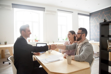 Happy millennial couple and smiling financial advisor, lawyer or realtor shaking hands at office...