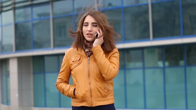 Beautiful young woman in yellow jacket is standing and talking on cell phone outdoor in the city