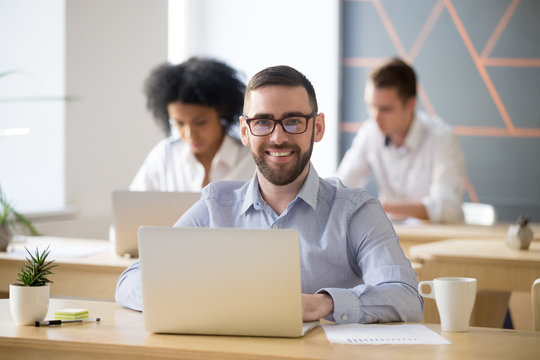 Smiling businessman in glasses sitting at desk with laptop in shared office, attractive millennial entrepreneur looking at camera in coworking, portrait of business professional posing at workplace
