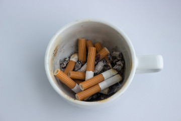 Cigarette butts with ashes in a white cup instead of ashtray on a light background. Dirty cup full of old cigarette and cigar butts. Unhealthy lifestle backdrop 