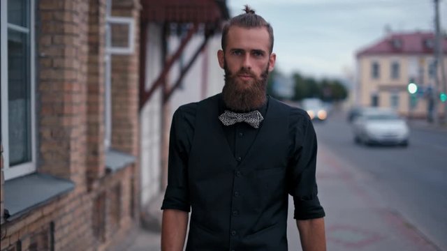 Young Trendy Bearded Man is Walking along the Street in the City and Looking to Camera