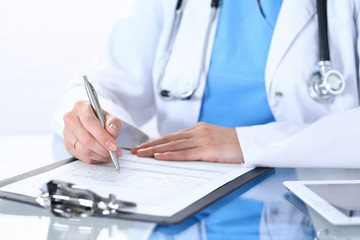 Female doctor filling up medical form by silver pen, closeup.  Physician sitting at the glass table in hospital office. Healthcare, insurance and medicine concept