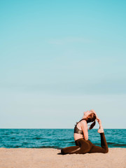 Young beautiful sporty woman in green clothing doing yoga asana on sea sandy beach near water. Girl practicing exercises. Health concept. Copy space.