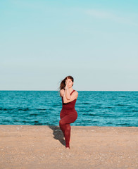 Young beautiful sporty woman in red costume doing yoga asana on sea beach near water. Girl practicing exercises. Health concept.
