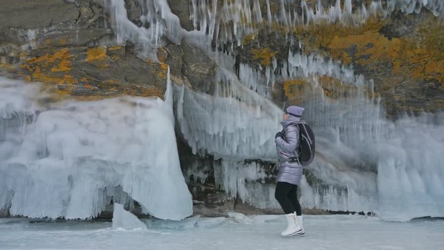 Travel of woman on ice of Lake Baikal. Trip to winter island. Girl is walking at foot of ice rocks. Traveler looks at beautiful ice grotto. Hiker wears sports glasses, silver jacket, backpack. Extreme