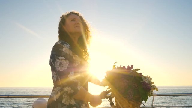 Young attractive woman in dress with vintage retro bicycle and lilac bouquet near sea during sunrise or sunset. Girl admires dawn and enjoys the moment of life on the coast of the ocean. 4k