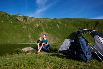 Fototapeta premium Romantic couple sits on rock near tent, backpack and trekking sticks and look into the distance against background of beautiful landscape of green mountains and lake and their foothills under blue sky