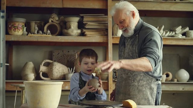 Caring silver-haired grandfather is teaching young cute grandson to work with clay on throwing-wheel in small workshop. Pottery, family hobby and handicraft concept.