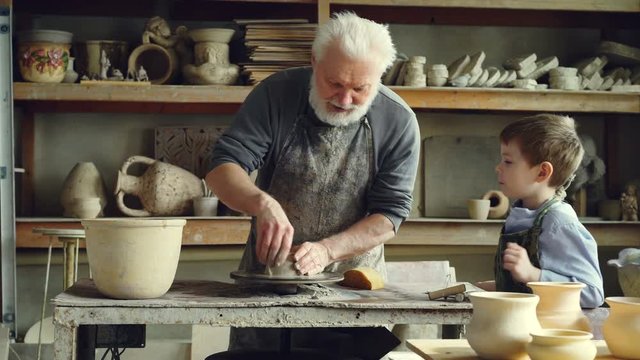 Caring senior grandfather is showing young grandson how to work with clay on throwing-wheel in small workshop. Pottery, family hobby and handicraft concept.