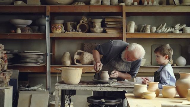 Senior ceramist is molding clay on spinning throwing-wheel while his little grandson is watching and learning. Sharing experience, family tradition and handwork concept.
