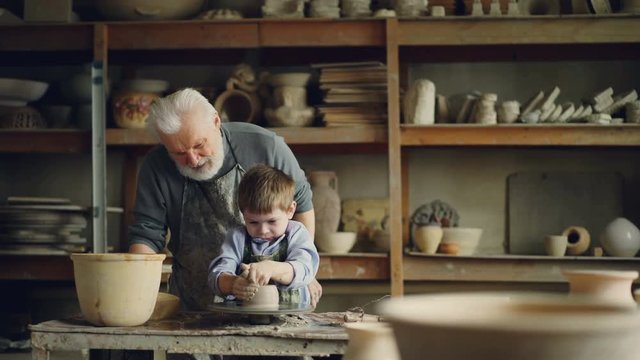 Caring grandfather experienced potter is teaching little boy how to work with clay on potter's wheel. Grandson is making mistake, patient grandpa is helping him.