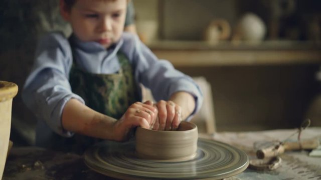 Serious boy is forming pot from clay on throwing wheel while attending pottery class in children's center. Interesting hobby, happy childhood and pottery concept.