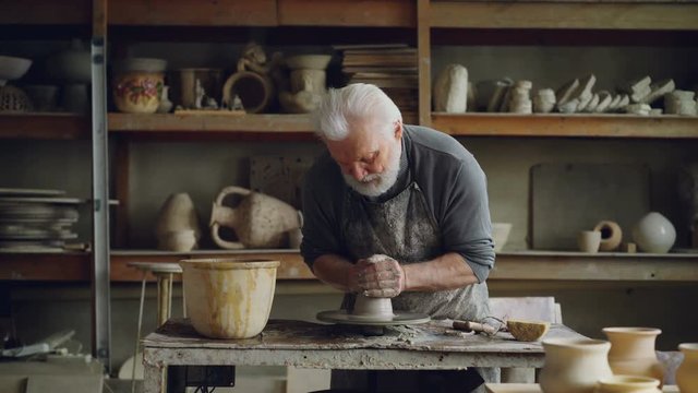 Hardworking grey-haired man is working with clay on potter's wheel, shaping piece of loam. Beautiful ceramic utensils, handmade pots and vases on shelves are visible.