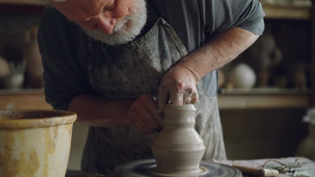 Hard-working senior potter is wetting hands in bowl with water and touching clay jar on spinning throwing-wheel. Bearded man is concentrated on work.