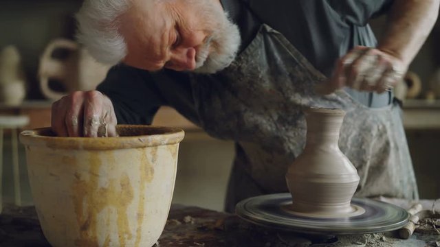 Elderly craftsman is creating ceramic jar on potter's wheel using professional tools. Workplace with handmade pots, beautiful vases and figures in background.