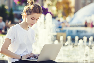 a beautiful teen woman is using her laptop computer outside, while sitting by a fountain in a town...