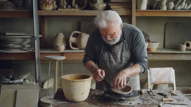Creative sculptor is making unusual broad bowl while standing at working table in his workshop. Bearded senior man is liiking at his clayware and appraising design and quality.
