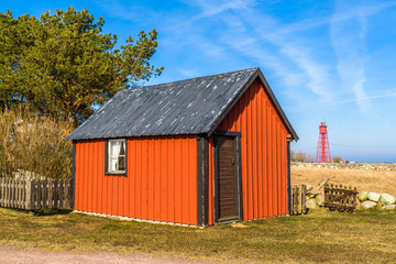 Obraz premium Kapelludden on Oland, Sweden. Weathered red wooden shed in coastal landscape with red lighthouse in background.
