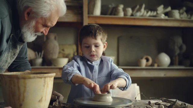 Little boy pottery learner is creating clayware on throwing-wheel while his experienced grandgather is talking to him and helping him. Making ceramics and family concept.